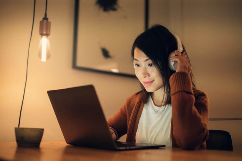 Medium shot of a young woman wearing headphones, studying online with laptop at home in the evening.