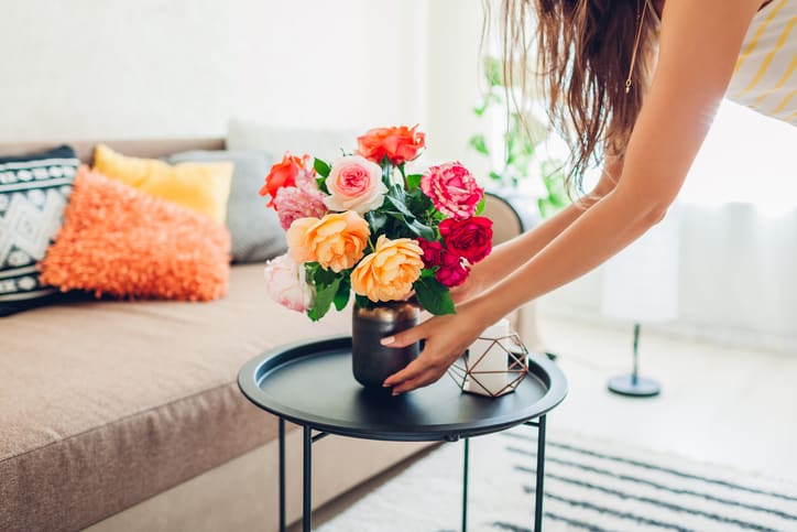 Woman puts vase with flowers roses on table. Housewife taking care of coziness in apartment. Interior and decor