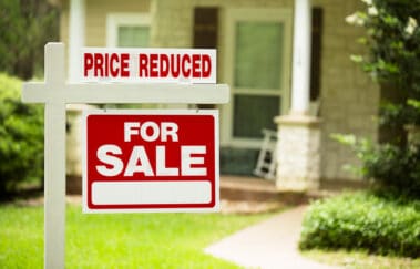 Red and white "Home for Sale, Price Reduced" sign in front of a stone, wood house that is for sale. Difficult listing concept.