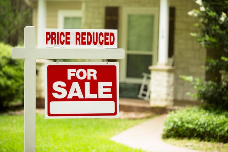 Red and white "Home for Sale, Price Reduced" sign in front of a stone, wood house that is for sale. Difficult listing concept.