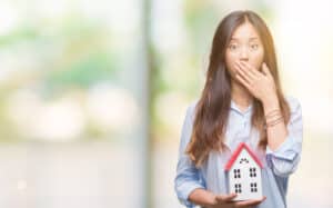 Young real estate broker holding house, covering mouth with hand, North Carolina real estate licensing pitfalls concept