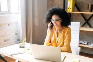 Charming young real estate broker talks on phone while working on laptop in modern office