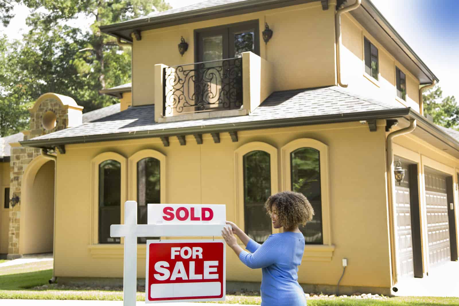 young adult realtor standing beside her for sale sign in front yard of home. She adds the SOLD sign and wears a blue top and jeans.